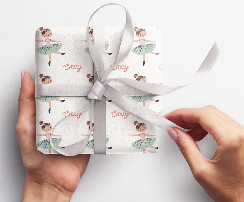 May include: White gift wrap with a repeating pattern of a ballerina in a green tutu. The name "Emily" is printed in pink on the wrapping paper.
