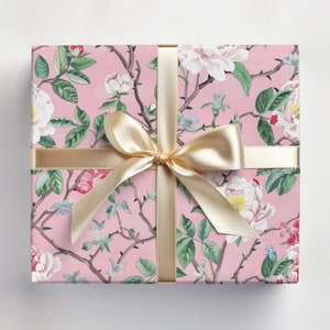May include: A wrapped gift box with a pink floral pattern featuring white and red flowers, green leaves, and brown branches. The box is tied with a gold satin ribbon and bow, creating a decorative and elegant presentation.