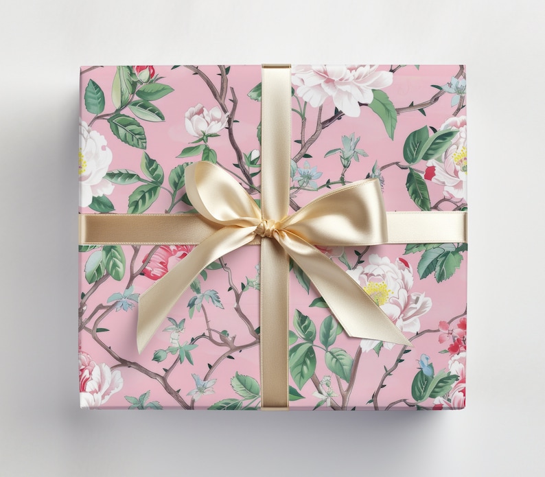 May include: A wrapped gift box with a pink floral pattern featuring white and red flowers, green leaves, and brown branches. The box is tied with a gold satin ribbon and bow, creating a decorative and elegant presentation.