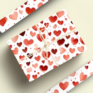 Op de afbeelding: Een cadeaudoos ingepakt in wit papier met een rood aquarel hartpatroon. De doos is vastgebonden met touw en heeft een rood hartpatroon aan de zijkanten. De doos wordt omringd door twee rollen cadeaupapier met hetzelfde patroon.