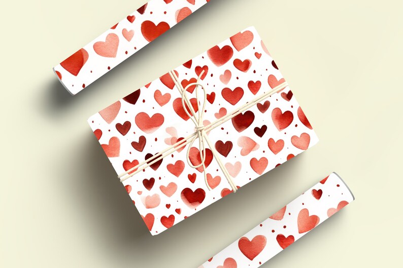 Op de afbeelding: Een cadeaudoos ingepakt in wit papier met een rood aquarel hartpatroon. De doos is vastgebonden met touw en heeft een rood hartpatroon aan de zijkanten. De doos wordt omringd door twee rollen cadeaupapier met hetzelfde patroon.