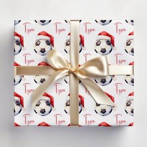 May include: White gift wrap with a repeating pattern of soccer balls wearing Santa hats. The name "Tyson" is repeated throughout the pattern. A gold ribbon is tied around the box.