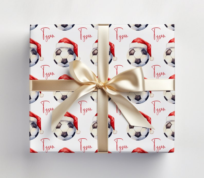 May include: White gift wrap with a repeating pattern of soccer balls wearing Santa hats. The name "Tyson" is repeated throughout the pattern. A gold ribbon is tied around the box.