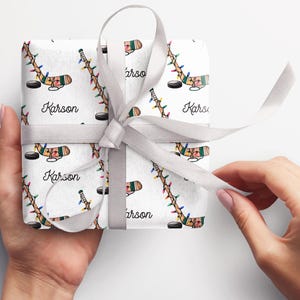 K&ouml;nnte beinhalten: Wei&szlig;e Geschenkpapier mit einem sich wiederholenden Muster aus mit Weihnachtslichtern umwickelten Hockeyschl&auml;gern und dem Namen "Karson". Das Geschenk ist mit einem wei&szlig;en Band gebunden.