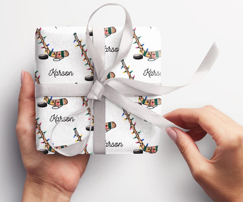 K&ouml;nnte beinhalten: Wei&szlig;e Geschenkpapier mit einem sich wiederholenden Muster aus mit Weihnachtslichtern umwickelten Hockeyschl&auml;gern und dem Namen "Karson". Das Geschenk ist mit einem wei&szlig;en Band gebunden.