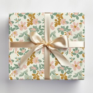 Op de afbeelding: Een cadeaudoos ingepakt in wit papier met een bloemenpatroon in roze, groene en gouden tinten. Een cr&egrave;mekleurig satijnen lint is bovenop de doos gebonden in een strik.