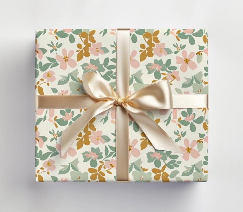 Op de afbeelding: Een cadeaudoos ingepakt in wit papier met een bloemenpatroon in roze, groene en gouden tinten. Een cr&egrave;mekleurig satijnen lint is bovenop de doos gebonden in een strik.
