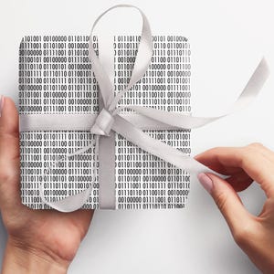 May include: A present wrapped in paper printed with a black and white binary code design, secured with a silver satin ribbon. The gift is held, with the ribbon being unwound.