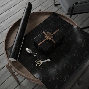 May include: A black gift box with a gold ribbon and bow, a roll of black wrapping paper, a pair of gold scissors, and a tape dispenser on a wooden table.