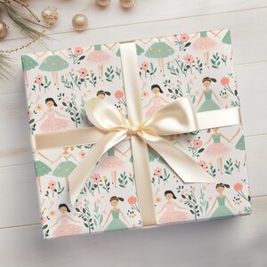 May include: White gift wrap with a repeating pattern of ballerinas in pink and green tutus dancing among pink and green flowers. A cream-colored satin ribbon is tied in a bow on top of the wrapping paper.