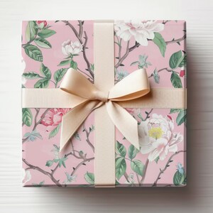 May include: A square gift box wrapped in pink floral patterned paper with a cream-colored ribbon and bow. The floral design features white and pink flowers, green leaves, and brown branches.