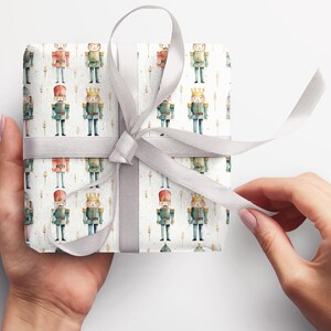 May include: A white gift box with a silver ribbon is wrapped in wrapping paper with a repeating pattern of watercolor nutcracker soldiers. The nutcracker soldiers are wearing blue and red uniforms with gold accents.