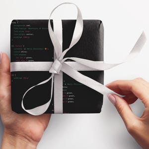 May include: A black gift box wrapped with a white ribbon and tied in a bow. The box is covered in code with the text "Merry Christmas!" in green and red.