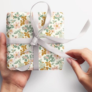 Op de afbeelding: Een cadeau verpakt in wit papier met een bloemenpatroon in roze, groene en gouden tinten. Het cadeau is vastgebonden met een wit satijnen lint.