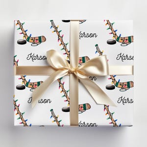 K&ouml;nnte beinhalten: Wei&szlig;e Geschenkpapier mit einem sich wiederholenden Muster aus Hockeyschl&auml;gern mit Weihnachtslichtern und dem aufgedruckten Namen "Karson". Das Geschenkpapier ist mit einem goldenen Band gebunden.
