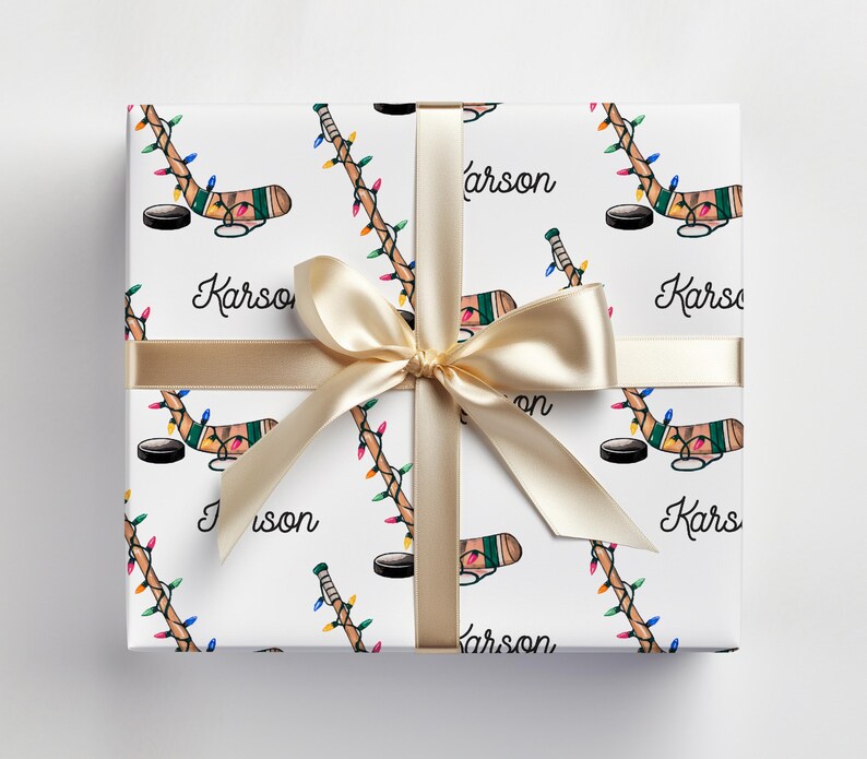 K&ouml;nnte beinhalten: Wei&szlig;e Geschenkpapier mit einem sich wiederholenden Muster aus Hockeyschl&auml;gern mit Weihnachtslichtern und dem aufgedruckten Namen "Karson". Das Geschenkpapier ist mit einem goldenen Band gebunden.