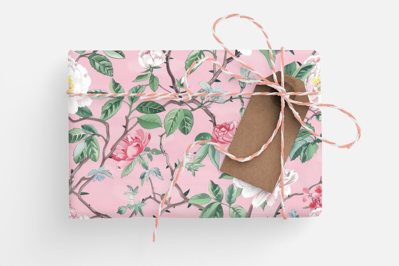 May include: A wrapped gift with a floral pattern featuring pink roses, white flowers, and green leaves on a pink background. The gift is tied with a white and peach twine and a brown tag.