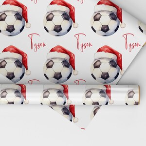 May include: White wrapping paper with a repeating pattern of soccer balls wearing Santa hats. The name "Tyson" is printed in red on the paper.
