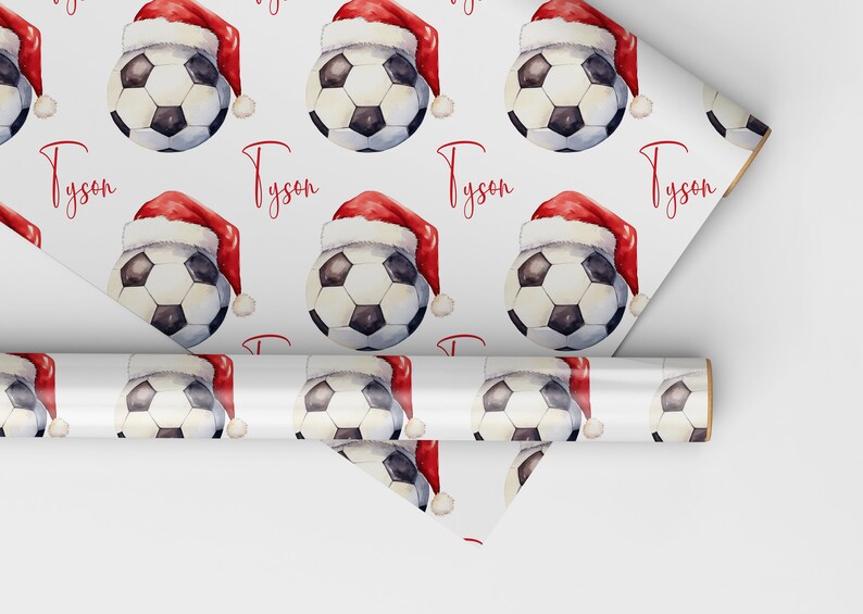 May include: White wrapping paper with a repeating pattern of soccer balls wearing Santa hats. The name "Tyson" is printed in red on the paper.