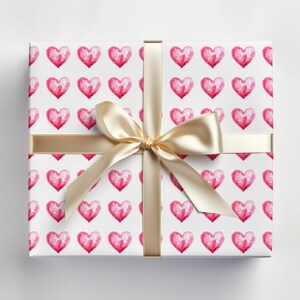 May include: A white gift box with a gold ribbon tied in a bow. The box is wrapped in pink watercolor heart patterned paper.