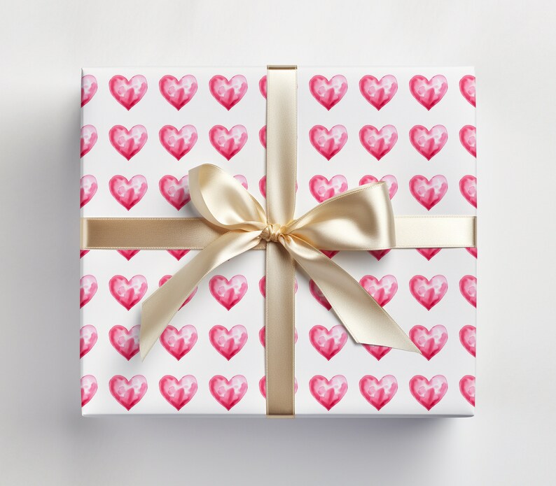 May include: A white gift box with a gold ribbon tied in a bow. The box is wrapped in pink watercolor heart patterned paper.