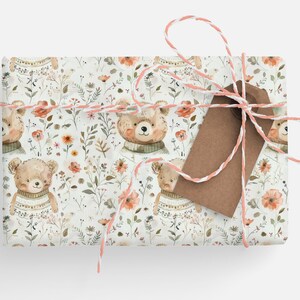 May include: A white gift wrap with a repeating pattern of brown bears wearing sweaters and pink flowers. The gift is tied with a white and pink twine and a brown tag.