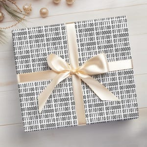 May include: A present wrapped in paper printed with binary code, tied with a cream satin ribbon and bow. Gold baubles are scattered around the gift on a light wooden surface.