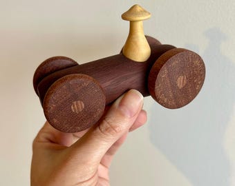 May include: A handcrafted wooden toy tractor with a dark brown body and light brown mushroom-shaped driver's seat. The wheels are circular and also dark brown. The toy is held in a person's hand against a plain white background.