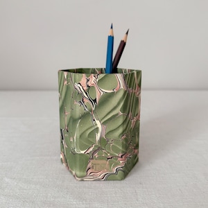 Può includere: Un portamatite esagonale con un motivo marmorizzato nei toni del verde salvia, bianco, nero e rosa tenue. L'esterno presenta una superficie strutturata e sfaccettata con un piccolo emblema color oro vicino alla base. Due matite sono all'interno.
