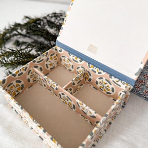 May include: Open jewelry box with a patterned design in shades of pink, yellow, and gray. The interior is divided into compartments. The lid is open, revealing a white interior and a blue trim. A sprig of greenery is in the background.