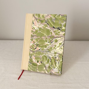 Op de afbeelding: Een hardcover boek met een gemarmerd patroon op de kaft in tinten olijfgroen, crème en zachtroze. De rug en de linkerkant zijn bekleed met een effen beige stof. Een dun rood lint als bladwijzer steekt aan de onderkant uit het boek.
