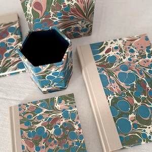 Könnte beinhalten: Eine Auswahl an Schreibwaren mit einem marmorierten Muster in Blau, Rosa und Grün. Das Set umfasst zwei Notizbücher, einen Stifthalter und eine kleine Box. Die Artikel haben cremefarbene Ränder. Der Stifthalter ist sechseckig.