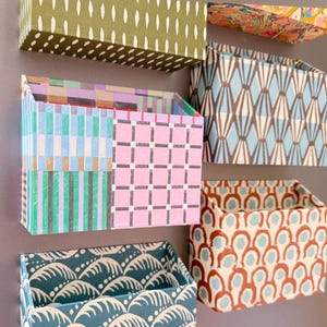 May include: A collection of five rectangular wall-mounted storage pockets featuring various patterns and colours, including olive green with white dashes, a pink grid, blue and green stripes, a teal wave print, and a brown and blue geometric design.