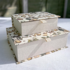 May include: Two rectangular storage boxes, one on top of the other. The lids display a pear and leaf pattern on a cream backdrop. The box sides are a light beige linen fabric. Each box has a small fabric tab for opening.