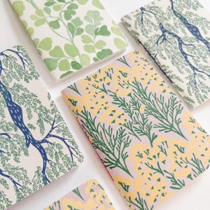 Könnte beinhalten: Eine Sammlung von Notizbüchern mit verschiedenen botanischen Mustern. Die Einbände zeigen Designs von grünen Blättern, Ästen und gelben Blumenarrangements. Die Notizbücher sind auf einer weißen Oberfläche angeordnet und präsentieren die verschiedenen Muster.