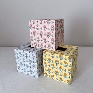 Può includere: Tre coperture quadrate per scatole di fazzoletti con un motivo floreale ripetuto. Una copertura è rosa chiaro con fiori rosa, una è azzurro con fiori blu e una è gialla con fiori arancioni. Ognuna presenta un'apertura rettangolare.