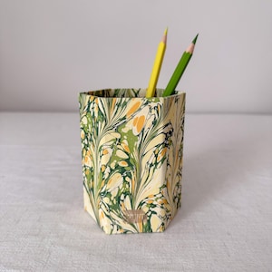 Puede incluir: Un portalápices hexagonal con un patrón jaspeado en tonos verde, amarillo y blanco. Dos lápices afilados, uno amarillo y otro verde, están colocados dentro del recipiente. La superficie exterior muestra un diseño fluido y arremolinado.