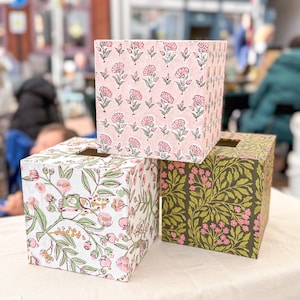 Könnte beinhalten: Drei dekorative Taschentuchbox-Hüllen. Eine ist hellrosa mit einem Blumenmuster, eine andere ist weiß mit rosa und grünen Blumendesigns, und die dritte ist dunkelgrün mit rosa Blumen und Blättern. Die Boxen sind quadratisch und stehen auf einer weißen Oberfläche.
