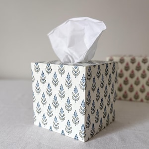 May include: A square tissue box with a white background and a repeating pattern of blue and grey leaves. White tissues protrude from the top opening. Other boxes with similar patterns are in the background.