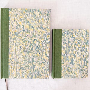 May include: Two hardback books of different sizes featuring a green fabric spine and a marbled paper cover. The marbled pattern consists of swirling designs in shades of sage green, pale yellow, white, and dark blue, creating a cohesive look.