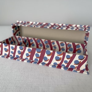 Könnte beinhalten: Geöffnete rechteckige Box mit Muster. Das Muster besteht aus diagonalen weinroten Streifen und blauen und orangefarbenen Kreisen auf cremefarbenem Hintergrund. Die Innenseite der Box ist cremefarben.