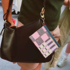 May include: A black leather shoulder bag with a gold-toned metal clasp and a small rectangular pouch attached to the strap. The pouch features a geometric pattern in shades of pink, purple, yellow, and dark gray, secured with a gold-toned clip.