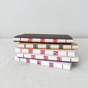 Peut inclure: Une pile de carnets faits à la main avec des coutures apparentes. Les couvertures sont de différentes couleurs, dont le marron, l'orange et le blanc. Les coutures sont rouges, bleues et noires, créant un motif décoratif.