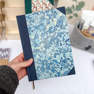 May include: A notebook with a marbled blue and white cover, and a navy blue spine and corners. A light brown ribbon bookmark is visible. Other stationery items are in the background. The notebook's cover has a unique pattern.