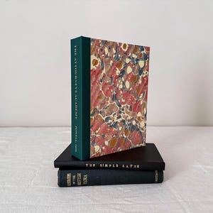 Op de afbeelding: Een stapel van drie boeken. Het bovenste boek heeft een donkergroene rug en een gemarmerde kaft met rode, witte en blauwe patronen, getiteld The Attorney's Academy. Daaronder liggen twee zwarte boeken, getiteld The Simple Layer en Illustrations of the British Flora.