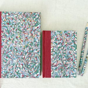 May include: Two hardcover journals and two pencils featuring a marbled pattern in shades of green, blue, white, and pink. Each journal has a solid red fabric spine. The pencils are wrapped in the same marbled paper as the journal covers.