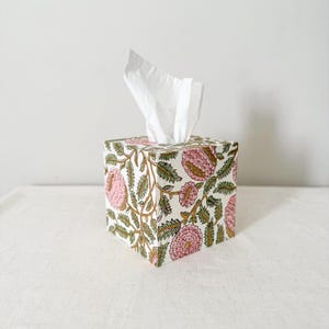 May include: A square tissue box featuring a floral design in pink, green, and gold on a white backdrop. White tissues protrude from the top. The box is placed on a white surface.