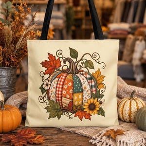 May include: A cream-colored tote bag with black handles featuring a central graphic of a patchwork pumpkin. The pumpkin is decorated with various patterns including plaid, floral, and polka dots, surrounded by autumn leaves and a sunflower.