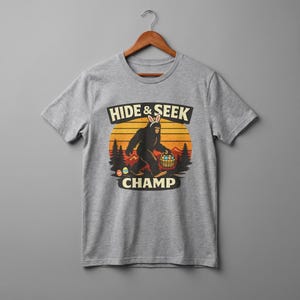 May include: A gray t-shirt featuring a graphic of Bigfoot wearing bunny ears and carrying a basket of Easter eggs. The text "HIDE & SEEK CHAMP" is above and below the graphic, set against a sunset backdrop.