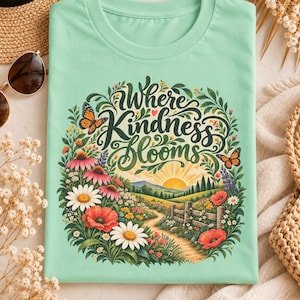 Könnte beinhalten: Ein helles mintgrünes T-Shirt mit einem farbenfrohen Grafikdruck einer Wiese mit Blumen, Schmetterlingen und einem Sonnenaufgang. Der Text "Where Kindness Blooms" steht in einer dunkelgrünen Schreibschrift über der Landschaft.